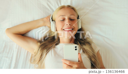 Happy relaxed middle-aged woman listening to music with phone in headphones lying on the bed Happy relaxed middle-aged woman listening to music with phone in headphones lying on the bed 136136466