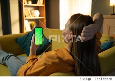 Young adult woman relaxing on a comfortable sofa at home, watching content on a green screen smartphone with ample room for custom creative application 136136626