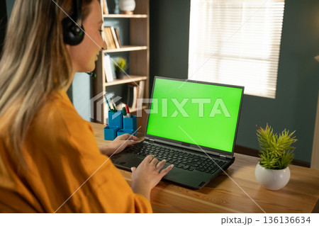 Young woman in her 20s wearing a headset and smiling while typing on a laptop with a green screen, working remotely from a bright home office and joining a virtual meeting Young woman in her 20s wearing a headset and smiling while typing on a laptop with a green screen, working remotely from a bright home office and joining a virtual meeting 136136634