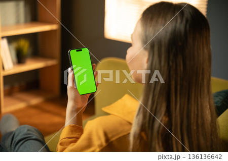 Young adult woman holding a modern smartphone with a blank green screen, relaxing on a comfortable sofa at home, providing ample space for custom content display 136136642