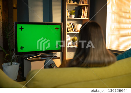 Woman relaxing on a couch at home in the evening, watching a television with a bright green screen and tracking markers, providing a customizable display for various media content 136136644