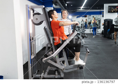 Woman exercising on chest press machine with personal trainer Woman exercising on chest press machine with personal trainer 136136797