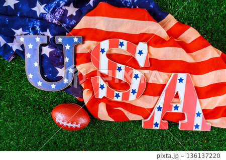 USA letters rest on flag with football nearby on grass, festive sports theme American pride. 136137220