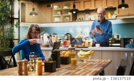 Senior adult browsing internet on smartphone and smiles in kitchen, relaxing with coffee and enjoying peaceful retired life. Retirement wellbeing, positive lifestyle and routine. Camera A. Senior adult browsing internet on smartphone and smiles in kitchen, relaxing with coffee and enjoying peaceful retired life. Retirement wellbeing, positive lifestyle and routine. Camera A. 136137510