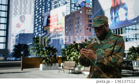 African american service person in full gear opens mobile phone map, walking through a modern downtown area in uniform. Soldier admiring skyscrapers and busy city street life. Camera A. African american service person in full gear opens mobile phone map, walking through a modern downtown area in uniform. Soldier admiring skyscrapers and busy city street life. Camera A. 136137512
