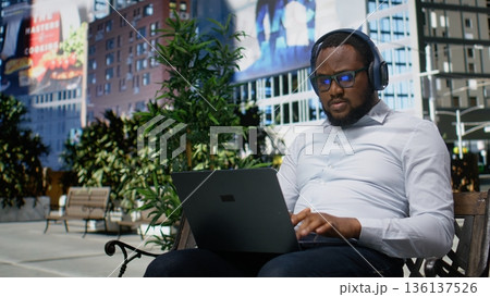 Confident adult man listening to focus music for remote work, sitting on a bench and using laptop for online tasks. Trendy activity in a metropolitan area, modern downtown avenue. Camera A. Confident adult man listening to focus music for remote work, sitting on a bench and using laptop for online tasks. Trendy activity in a metropolitan area, modern downtown avenue. Camera A. 136137526