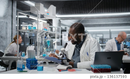 Lab researcher looking through scientific microscope lens to study cells. African american man performing biological sample examination with high tech optical equipment, camera A Lab researcher looking through scientific microscope lens to study cells. African american man performing biological sample examination with high tech optical equipment, camera A 136137593