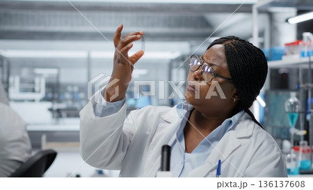 Female molecular biologist studying genetic tissue sample on glass slide under magnification. African american woman searching for mutation patterns using advanced optical equipment, camera B 136137608