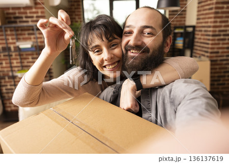 Excited young couple posing with house keys after moving into new brick wall apartment. Smiling caucasian man and woman hugging, holding cardboard box, celebrating first home together. 136137619