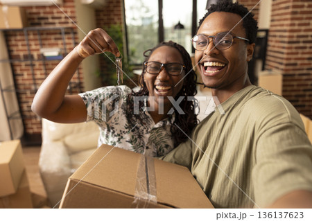 Cheerful African American spouses smile at camera while holding house keys and moving boxes. Happy male and female partners celebrate new home with laughter and selfie in modern living room. Cheerful African American spouses smile at camera while holding house keys and moving boxes. Happy male and female partners celebrate new home with laughter and selfie in modern living room. 136137623