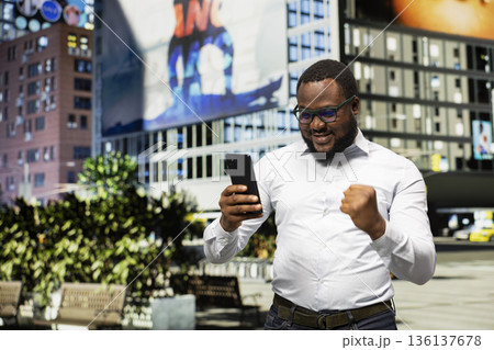 African american person being joyous and satisfied with good news on smartphone, walking on a street downtown and enjoying achievement. Man excited about career accomplishment. 136137678