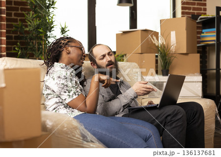 Black woman relaxing on sofa with white husband, surrounded by boxes, browsing laptop for furniture and home ideas. Young couple shops online and explores neighborhood information in new apartment. 136137681