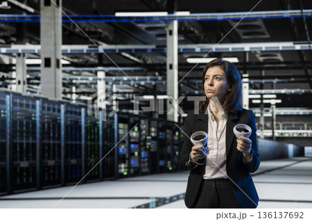 Server room manager using VR equipment to do maintenance on storage infrastructure rigs. Female engineer uses virtual reality gear to inspect high tech data center electronics 136137692