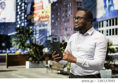 Carefree casual guy strolling downtown and browsing tablet near busy avenue, taking a relaxed walk on city streets. Modern architecture, urban culture and the vibrant pace of city life. 136137693