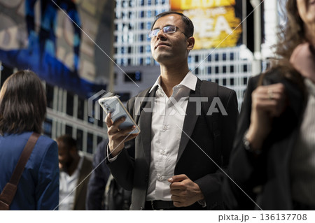Indian male modern professional checking apps on phone at red light, standing in the street surrounded by pedestrians crowd. Morning commute of businessman walking to the office. 136137708