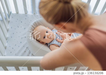 Mother gently sitting beside the crib and stroking her baby resting in a cozy cocoon nest. Tender intimate moment of care and trust at home. Motherhood, infant sleep and emotional bonding concept 136138206