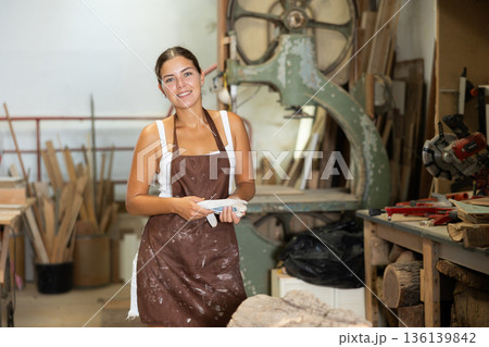 Girl employee of workshop cuts wooden board into parts using saw Girl employee of workshop cuts wooden board into parts using saw 136139842