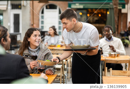 On open area of cafe, guy waiter serves cafe establishments girl visitor, puts food on table. 136139905