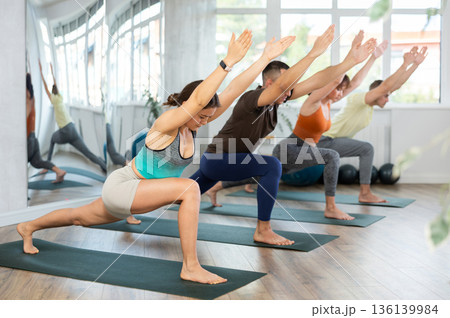 Lovers of active lifestyle on yoga mat perform doing version of warrior pose in gym Lovers of active lifestyle on yoga mat perform doing version of warrior pose in gym 136139984