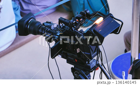 Professional video camera on a tripod with lens and screen, recording at an event 136140145
