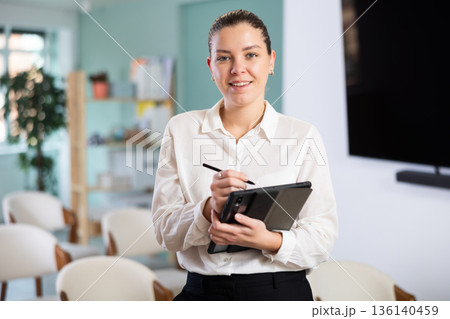 Positive friendly young girl secretary with digital tablet in hands gesture inviting visitors to enter in office 136140459