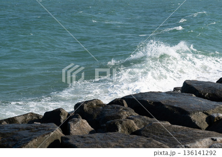 Powerful sea waves crash against dark rock formation creating white foam splash on blue ocean water near nature coast landscape outdoor view Powerful sea waves crash against dark rock formation creating white foam splash on blue ocean water near nature coast landscape outdoor view 136141292