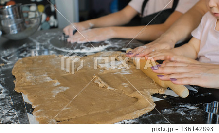 Mother helps her young daughter press and smooth ginger cookie dough on the kitchen table while the older daughter assists nearby 136142893