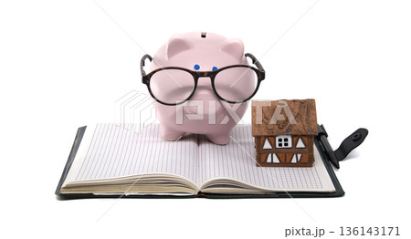 Smart piggy bank with glasses and house model on open notebook, symbolizing financial planning Smart piggy bank with glasses and house model on open notebook, symbolizing financial planning 136143171