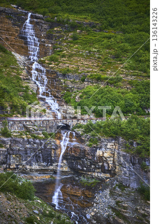 Waterfall Cascading Over Rocky Cliff with Scenic Mountain Road and Lush Greenery 136143226