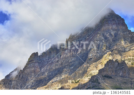 Majestic Rocky Mountain Peak with Dramatic Cloud Cover and Sunlit Cliff Faces Majestic Rocky Mountain Peak with Dramatic Cloud Cover and Sunlit Cliff Faces 136143581