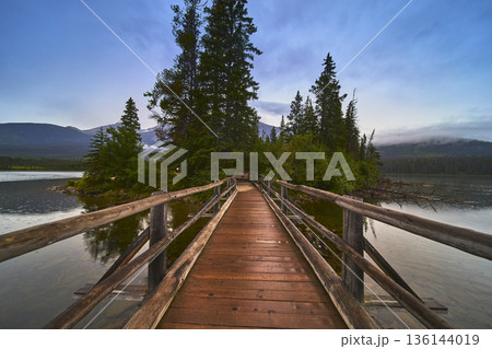 Wooden Bridge Leading to Forested Island on Tranquil Lake with Mountain Backdrop 136144019