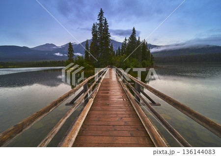 Bridge Leading to Forested Island on Calm Lake with Mountain Backdrop at Dusk 136144083