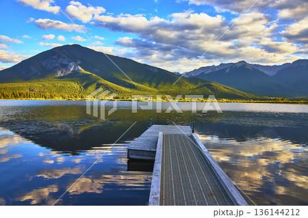 Dock Leading to Tranquil Lake with Mountain Reflections and Dramatic Sky Dock Leading to Tranquil Lake with Mountain Reflections and Dramatic Sky 136144212