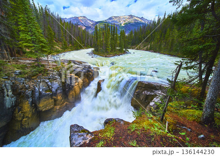 Waterfall Flowing Over Rocky Cliff Surrounded By Pine Trees And Majestic Mountain Peaks 136144230