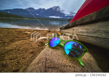 Sunglasses on Rocky Shore with Canoe and Mountain Reflections at Lakeside 136144282