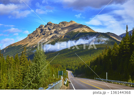 Highway Leading Into Mountain With Cloud and Evergreen Forest in Vibrant Daylight 136144368