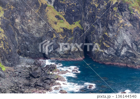 Rugged dark volcanic sea cliff wall with natural cave entrance and deep blue ocean water crashing onto rocky shoreline. Rugged dark volcanic sea cliff wall with natural cave entrance and deep blue ocean water crashing onto rocky shoreline. 136144540