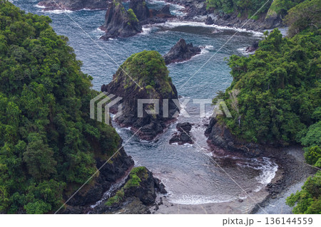 Overhead view of lush green forested cliffs and dark volcanic rock formations in a secluded bay with turquoise ocean water. Overhead view of lush green forested cliffs and dark volcanic rock formations in a secluded bay with turquoise ocean water. 136144559