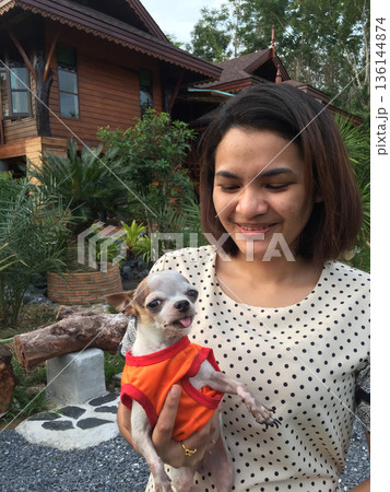 Asian woman holding small chihuahua dog outdoors front wood house 136144874