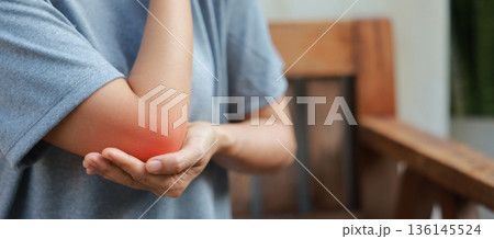 Close-up of woman hand holding painful injured elbow with red inflamed spot. Concept of joint bone pain, muscle injury, arthritis, tendonitis, or ache from sports or work in outdoor background. 136145524