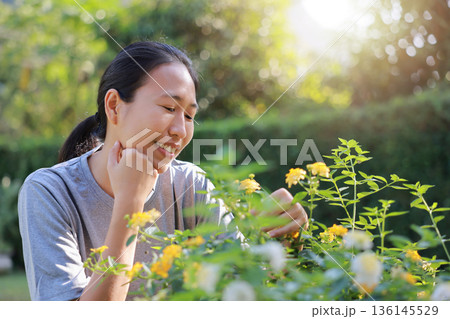 Close-up of a happy Asian woman smiling and enjoying purple flowers in her green garden. Beautiful female portrait in sunny backyard park. Concept of nature lovers, wellness, and outdoor lifestyle. Close-up of a happy Asian woman smiling and enjoying purple flowers in her green garden. Beautiful female portrait in sunny backyard park. Concept of nature lovers, wellness, and outdoor lifestyle. 136145529