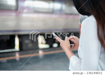 A woman checks her smartphone while waiting at a train station, with a blurred train passing by in the background. 136149631