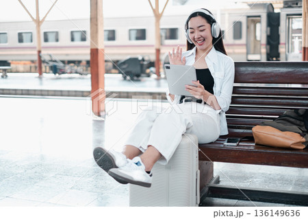 Young woman with headphones using a tablet at a train station, sitting on luggage, smiling and waving. 136149636