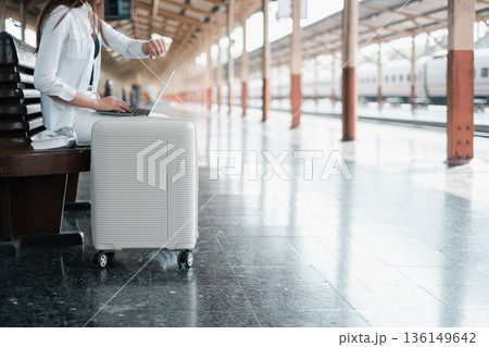 Person using a laptop while sitting on a suitcase at a train station, symbolizing travel and technology. Person using a laptop while sitting on a suitcase at a train station, symbolizing travel and technology. 136149642