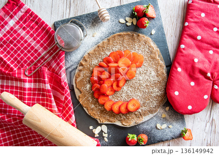 Homemade galette pie cake with strawberries and almond, placed on black desk and wooden table. Red towel with cooking tools on the side 136149942