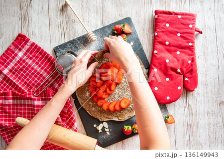 Galette with strawberries preparation, detail of hands of cooker. Pie cake placed on black desk and wooden table with cooking tools on the side. 136149943