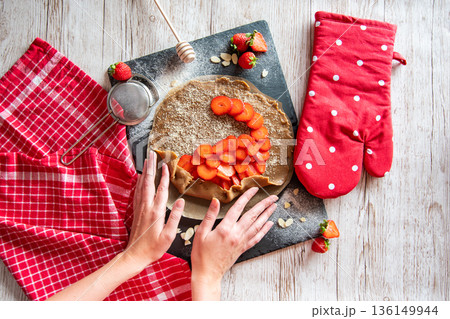 Galette with strawberries preparation, detail of hands of cooker. Pie cake placed on black desk and wooden table with cooking tools on the side. 136149944