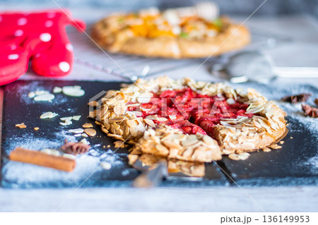Homemade galette pie cake with strawberries and almond, placed on black desk and wooden table. Red towel with cooking tools on the side Homemade galette pie cake with strawberries and almond, placed on black desk and wooden table. Red towel with cooking tools on the side 136149953