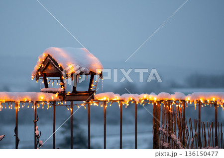 Wooden birdhouse covered by snow, with glowing christmas lights placed in small roof garden. Wooden birdhouse covered by snow, with glowing christmas lights placed in small roof garden. 136150477