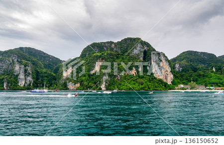 Panoramic wide-angle view of Ko Phi Phi island in Thailand from the sea, surrounded by numerous small and large boats on the clear tropical water under a sunny sky. 136150652